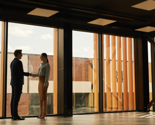 Wide Angle View Of Real Estate Agent Shaking Hands With Client While Standing In Empty Office Building Interior Lit By Sunlight